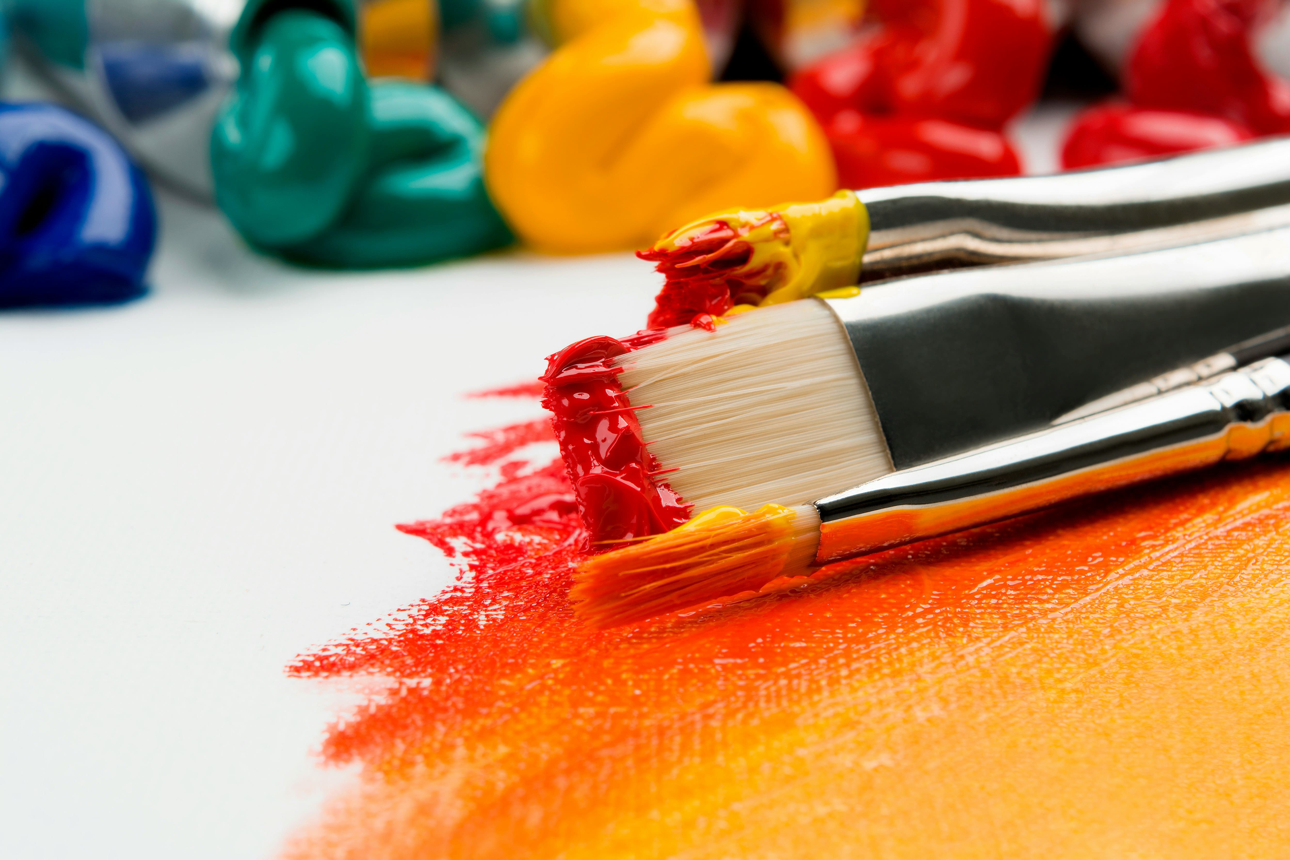 Symbolbild für Konstruktivismus: drei Pinsel mit orangener und gelber Farbe liegen auf einer Leinwand, die bereits zum Teil in diesen Farben bemalt wurde. Im Hintergrund laufen Farbtuben in verschiedenen Farben aus.