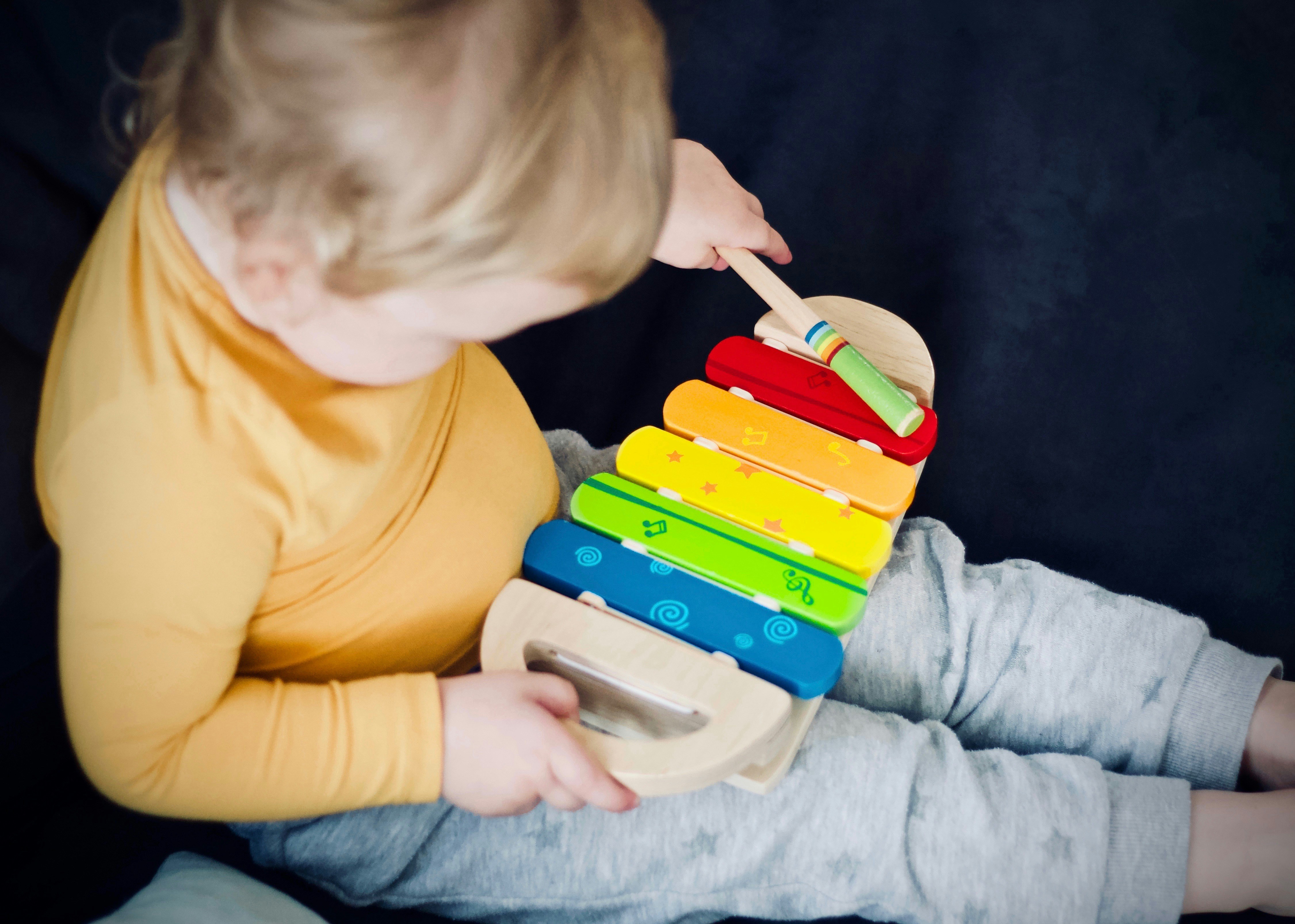 Symbolbild für Konstruktivistisches Lernen: Kleinkind sitzt auf Couch und schlägt mit einem Holzstick auf einem bunten Xylophon herum, lernt also durch eigenes Experimentieren.